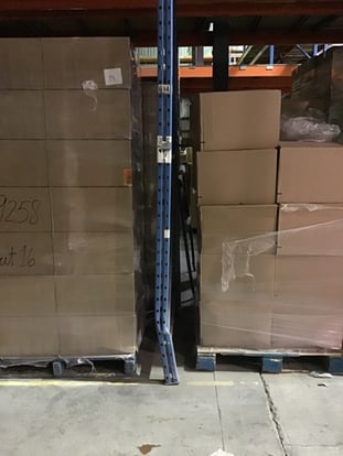 Blue pallet rack upright bent out of alignment near the base from forklift impact, still supporting a loaded bay