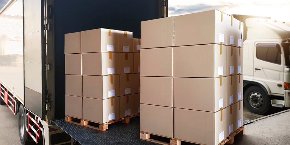 Stacks of cardboard boxes on pallets being loaded into a truck at a warehouse dock, illustrating cross-docking logistics with minimal storage and fast outbound flow.