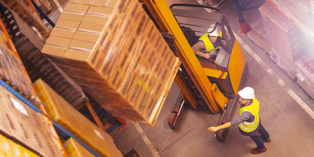 Two warehouse workers placing pallets onto pallet racks, illustrating safe material handling and rack use.