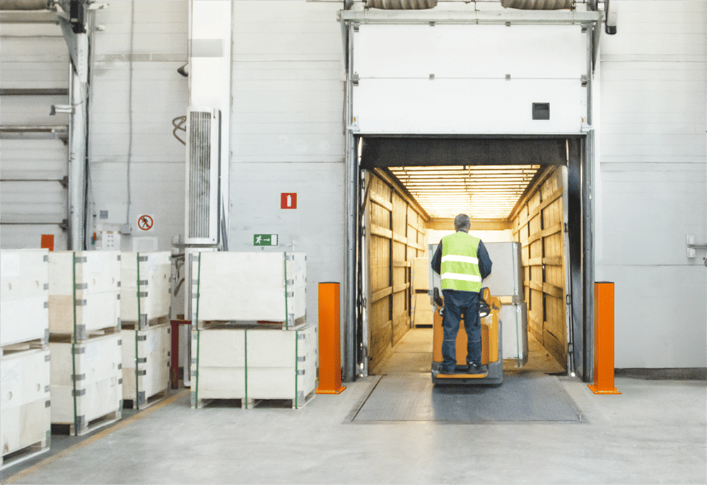 Two orange rectangle safety DAMO Bollards anchored in front of a warehouse docking door while a truck gets loaded with cargo