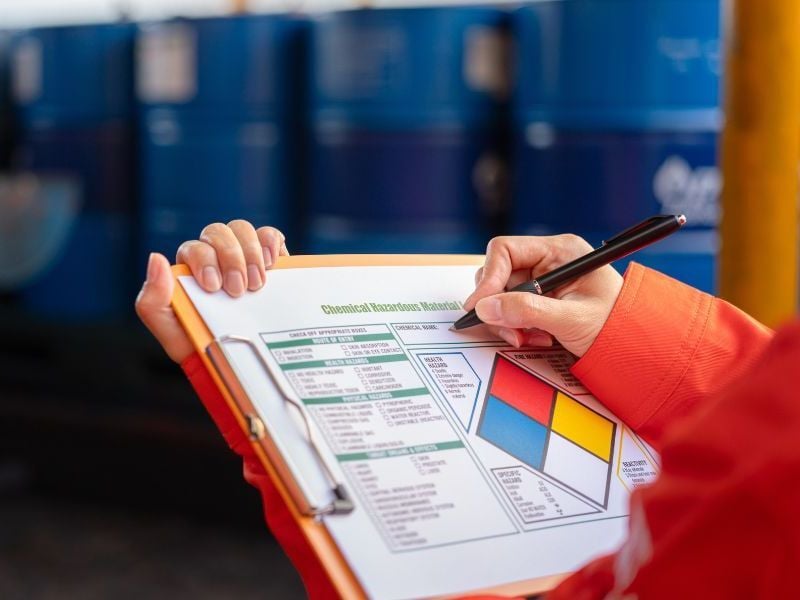 Worker filling out a chemical hazard label and safety checklist on a clipboard in front of stacked blue chemical drums in a warehouse.