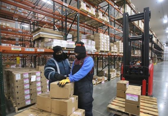 Two workers wearing insulated PPE and balaclavas handle boxes in a freezer warehouse aisle with pallet racking and a forklift nearby.