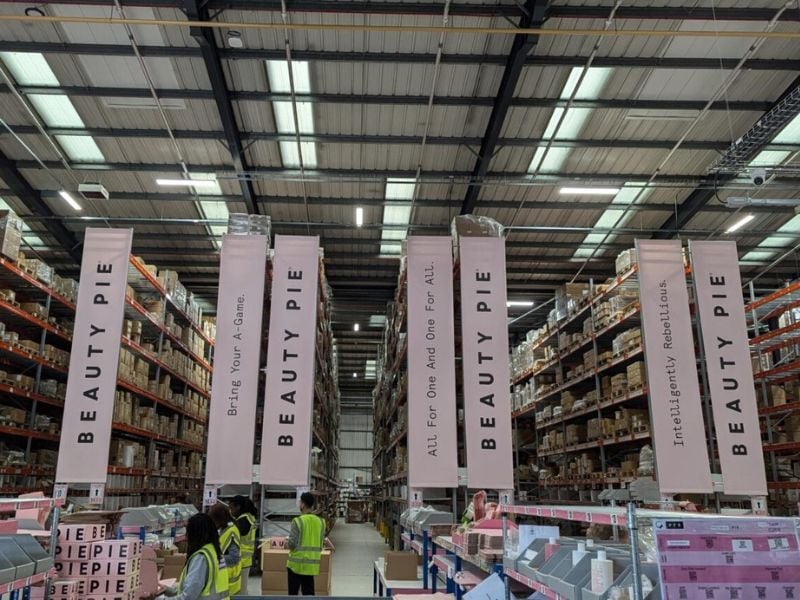 Intérieur d’un entrepôt de produits cosmétiques avec des palettiers hauts, des bannières de marque Beauty Pie et des employés en gilets de sécurité gérant la préparation des commandes.
