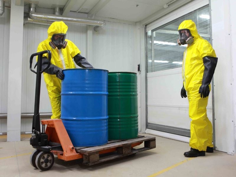 Two workers in yellow hazmat suits and respirators handling blue and green chemical drums on a pallet jack inside a warehouse.