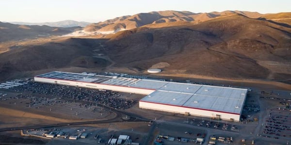 Aerial view of Tesla’s Gigafactory Nevada, showcasing the sprawling industrial facility set in the desert landscape with solar panels on the roof and surrounding infrastructure.