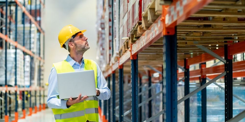 Warehouse inspector looking at pallet rack beams with laptop for safety and compliance