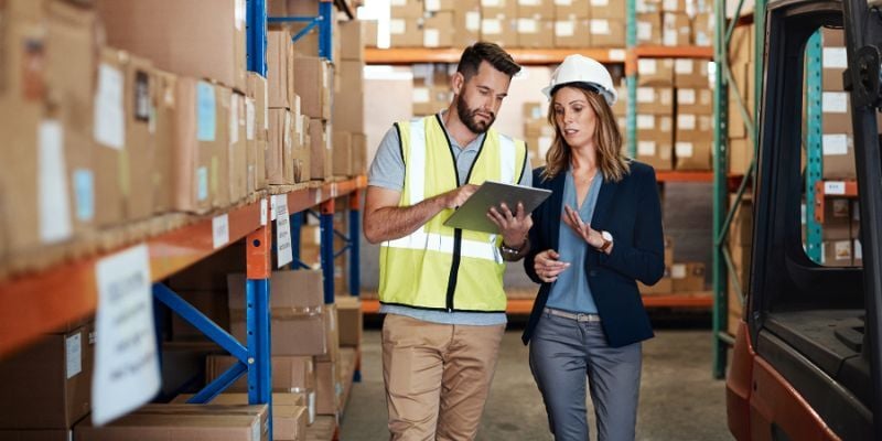 Warehouse manager reviewing pallet rack inspection results on a tablet with a warehouse worker beside storage racking