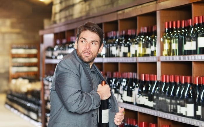Man awkwardly attempting to steal a bottle of wine in plain sight, glancing nervously over his shoulder in a store aisle filled with bottles.