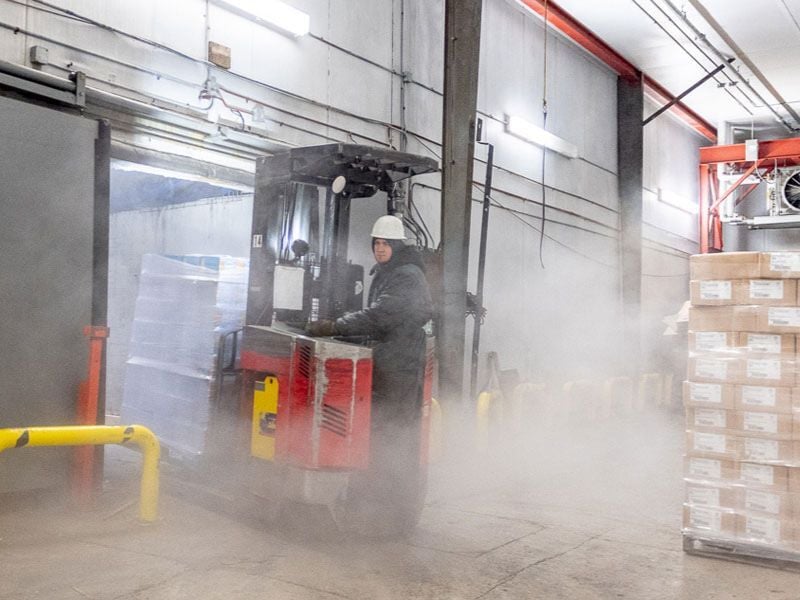 Cariste portant un EPI pour temps froid et un casque de sécurité, conduisant dans une zone de chargement d’entrepôt congélateur embrumée avec condensation visible.
