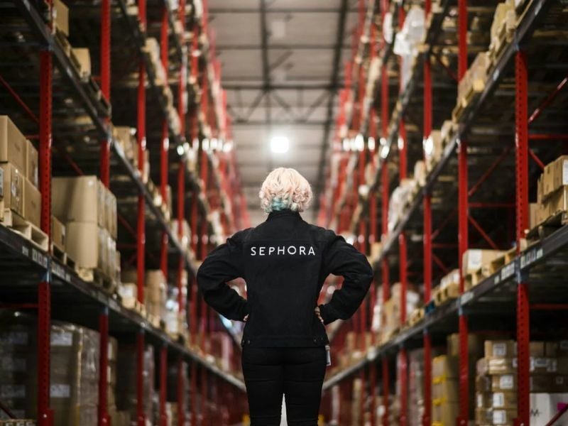 Employé de Sephora debout entre de hauts palettiers rouges dans un entrepôt, face à l’avant, les mains sur les hanches, inspectant l’environnement de stockage.